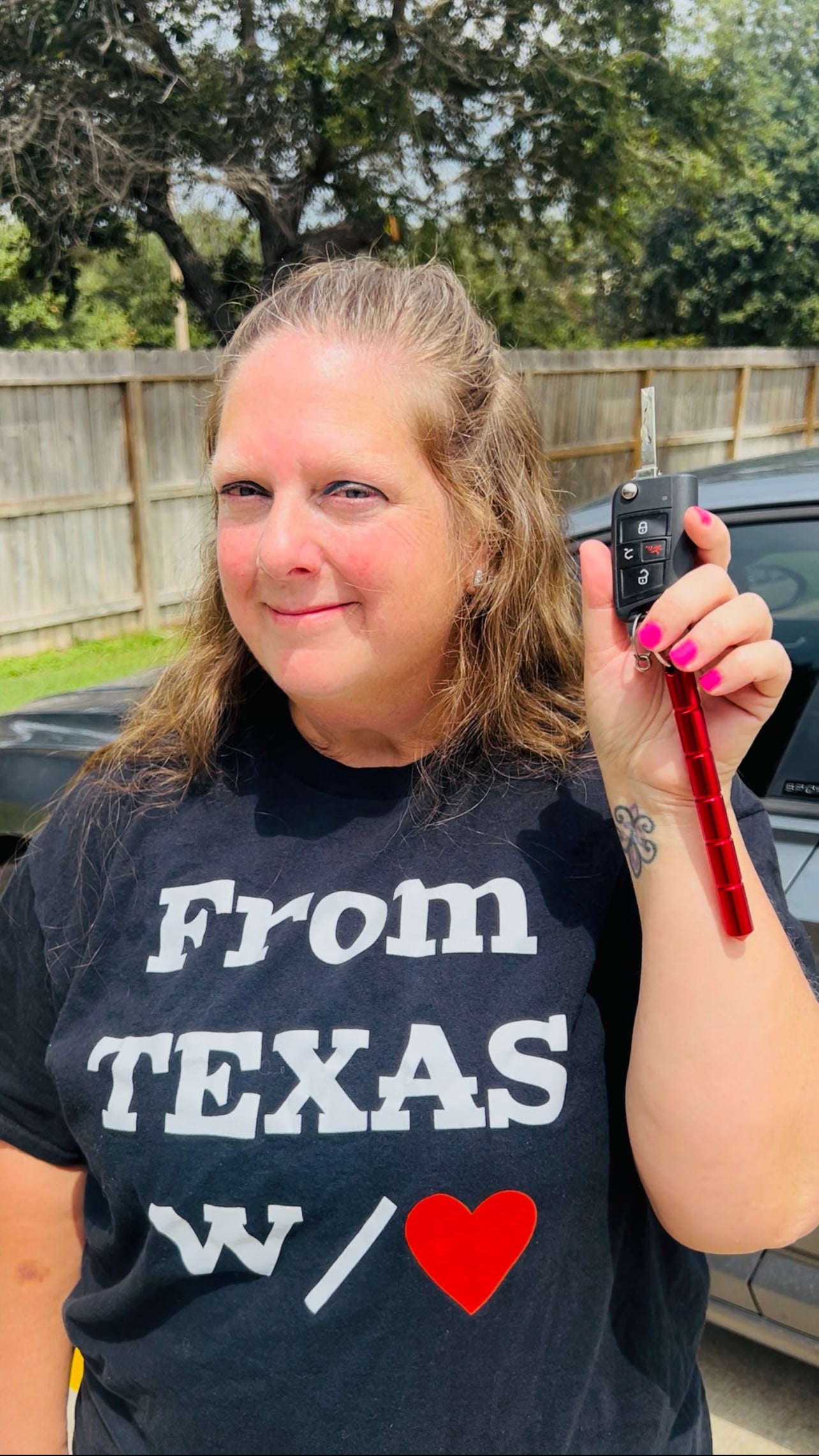 Woman wearing From Texas W/❤️ shirt holding red self-defense keychain with car keys outside.