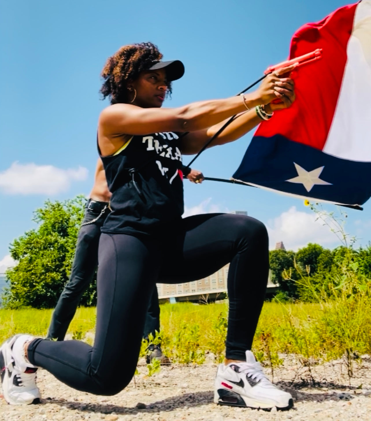 Woman of color wearing a hat kneeling outdoors holding GRUPS® Grip Trainer with Texas flag, symbolizing strength, safety, self-defense training and empowerment.